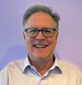 Smiling middle-aged man with gray hair, black-rimmed glasses, and a white shirt in front of a purple background.